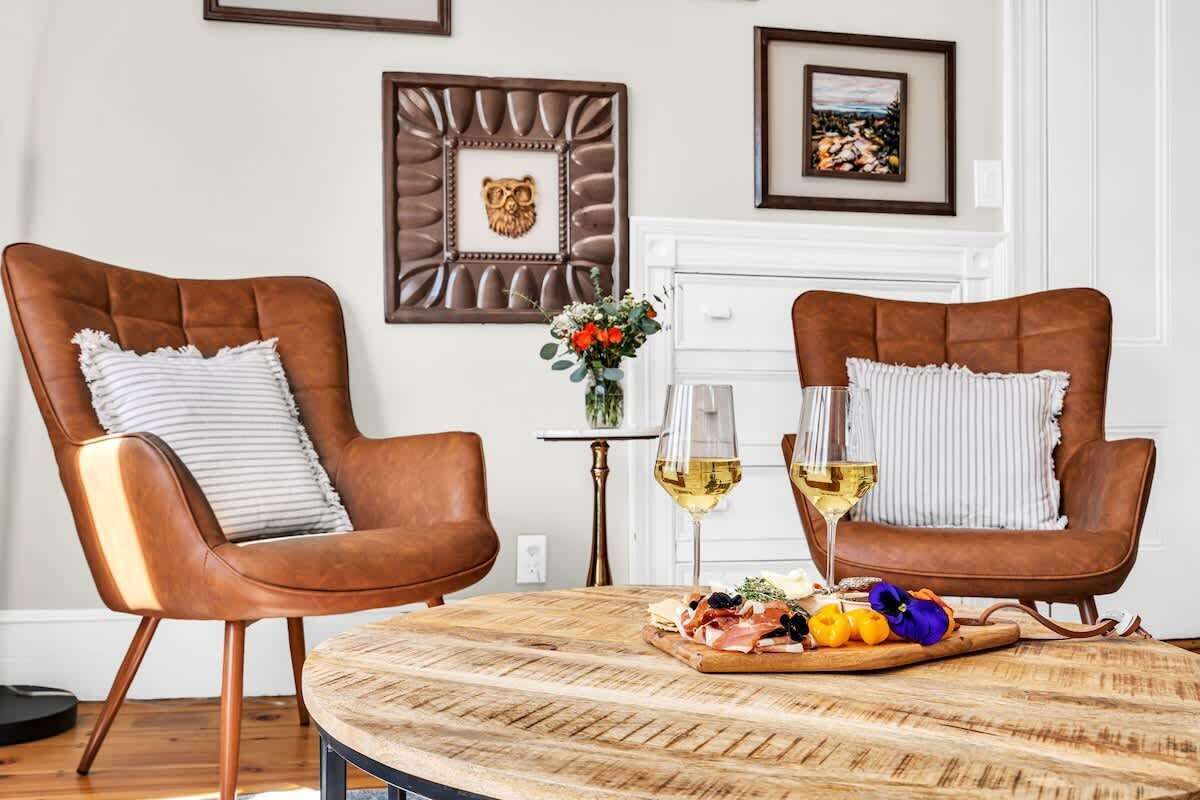 A dining area with two brown leather chairs, a round wooden table, and a small side table with a vase and two wine glasses.