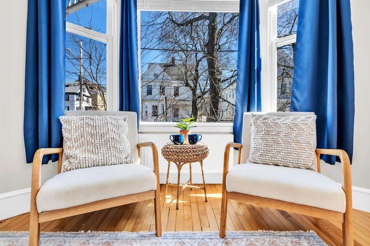 Un coin salon confortable avec deux fauteuils et une petite table, une fenêtre avec des rideaux bleus et une vue sur une maison et des arbres.