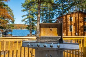 A portable grill with four burners, a chimney, and a control panel, set on a wooden deck with a view of a lake and a cabin in the background.