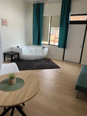 Una habitación moderna con suelo de madera, una mesa redonda con una planta, un tapete negro, una silla blanca, un sofá color turquesa, una bañera y una puerta con un cartel.