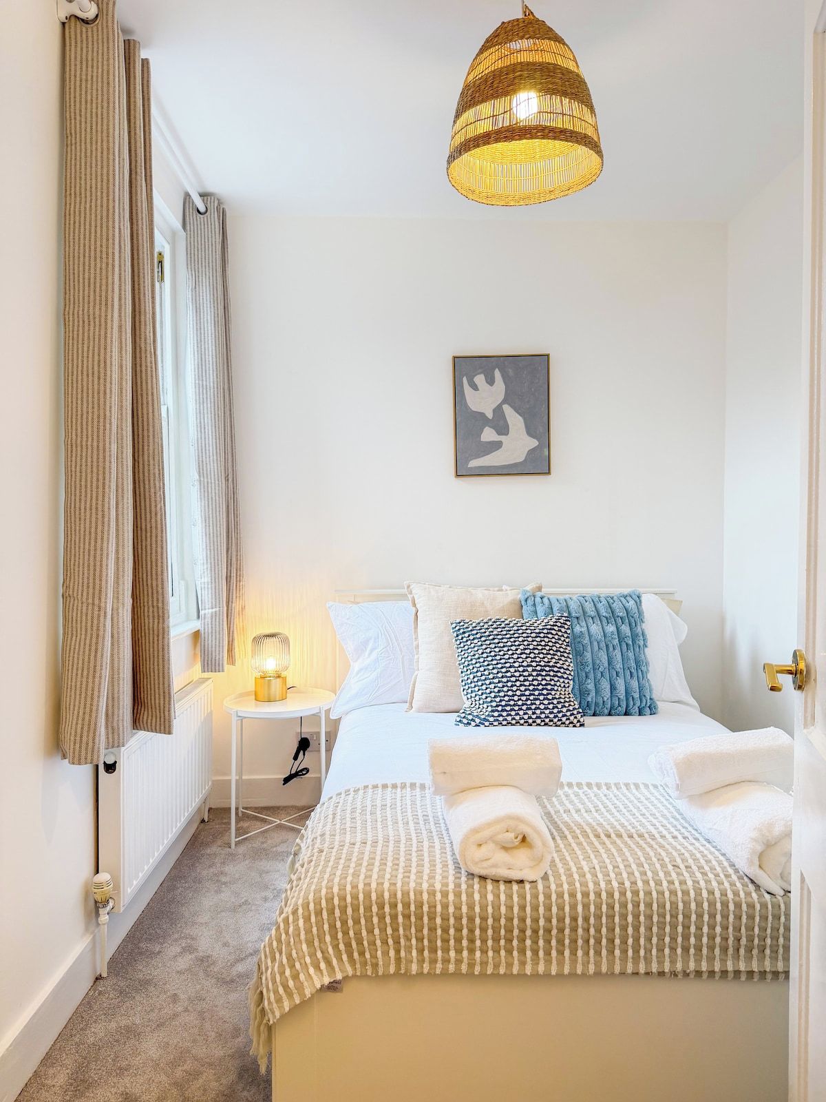 A bedroom with a bed, a bedside table with a lamp, a hanging light fixture, and a framed artwork on the wall.