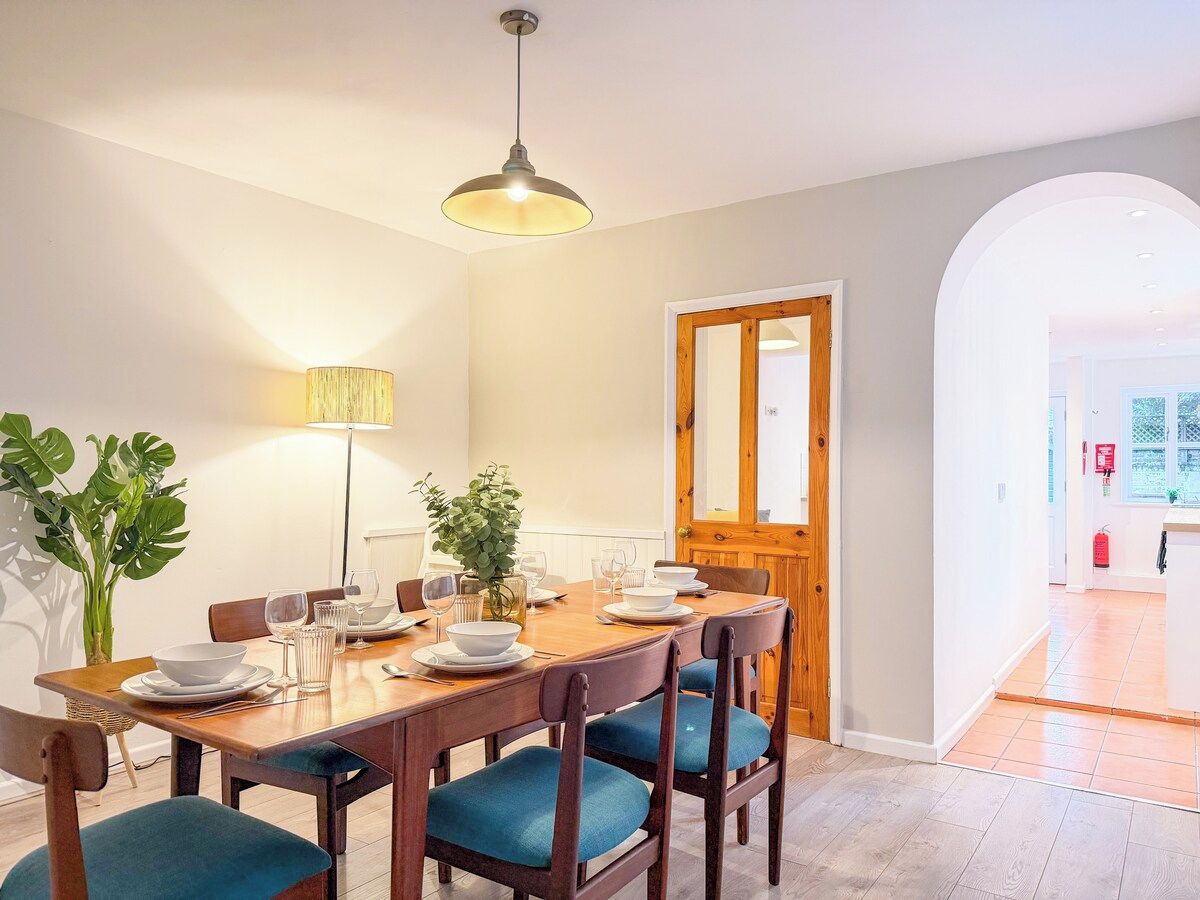 Un coin repas avec une table en bois dressée pour quatre personnes, des chaises à l’assise rembourrée en tissu bleu, un luminaire suspendu et une plante en pot.