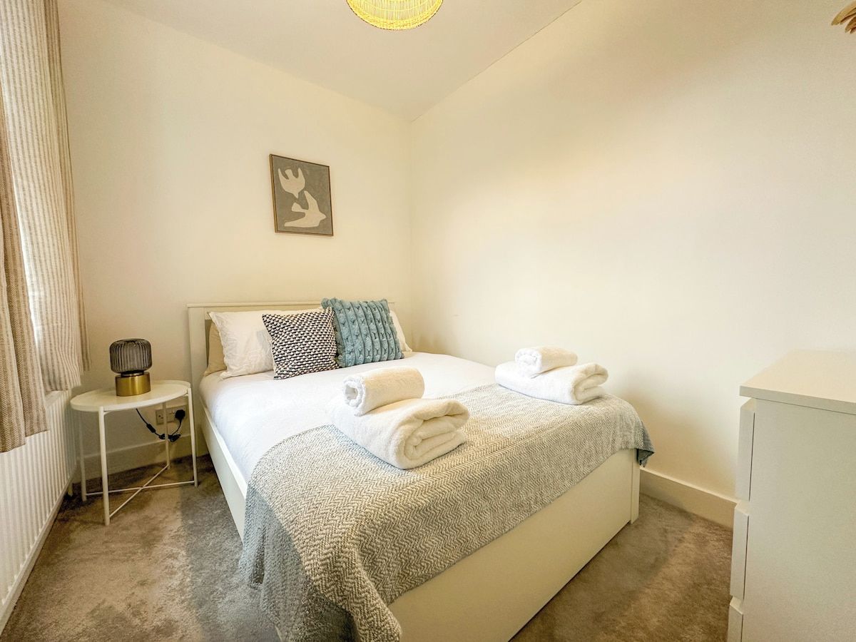 A hotel room with a bed, a bedside table with a lamp, a wall-mounted artwork, and a small cabinet.