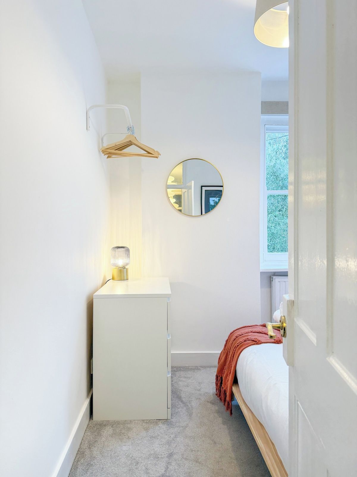 A small, compact bedroom with a single bed, a bedside table, a mirror, and a window.