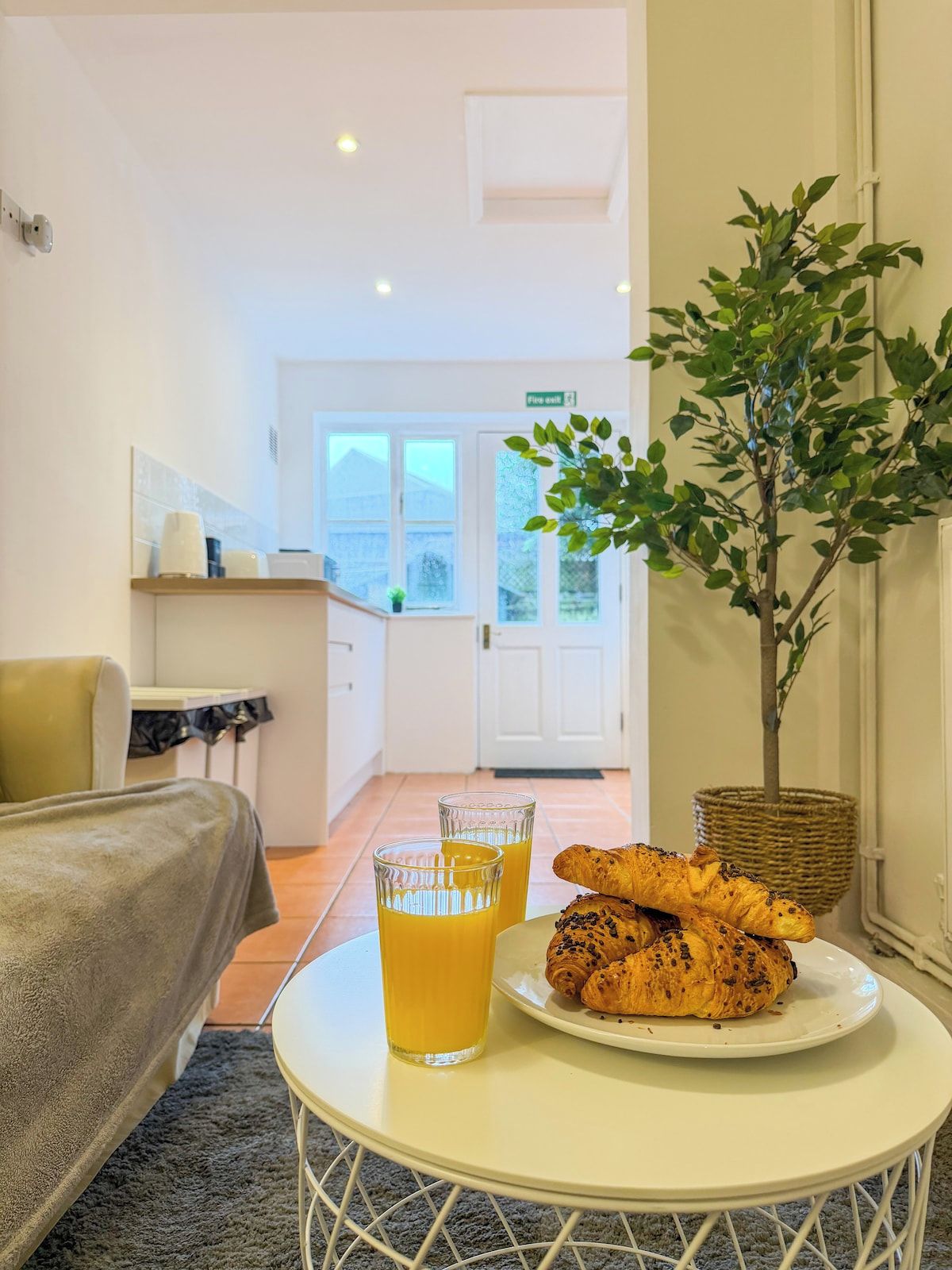 A hotel room with a bed, a small round table with a plate of croissants and glasses of orange juice, a potted plant, and a view of the kitchen and outside.