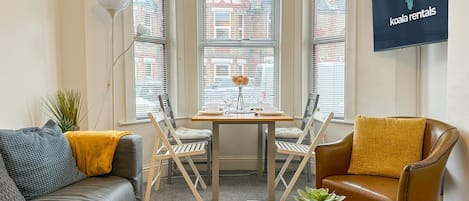 Una sala moderna con un sofá, una silla, una mesita de centro y una mesa de comedor. Hay una planta sobre la mesita y un televisor montado en la pared.