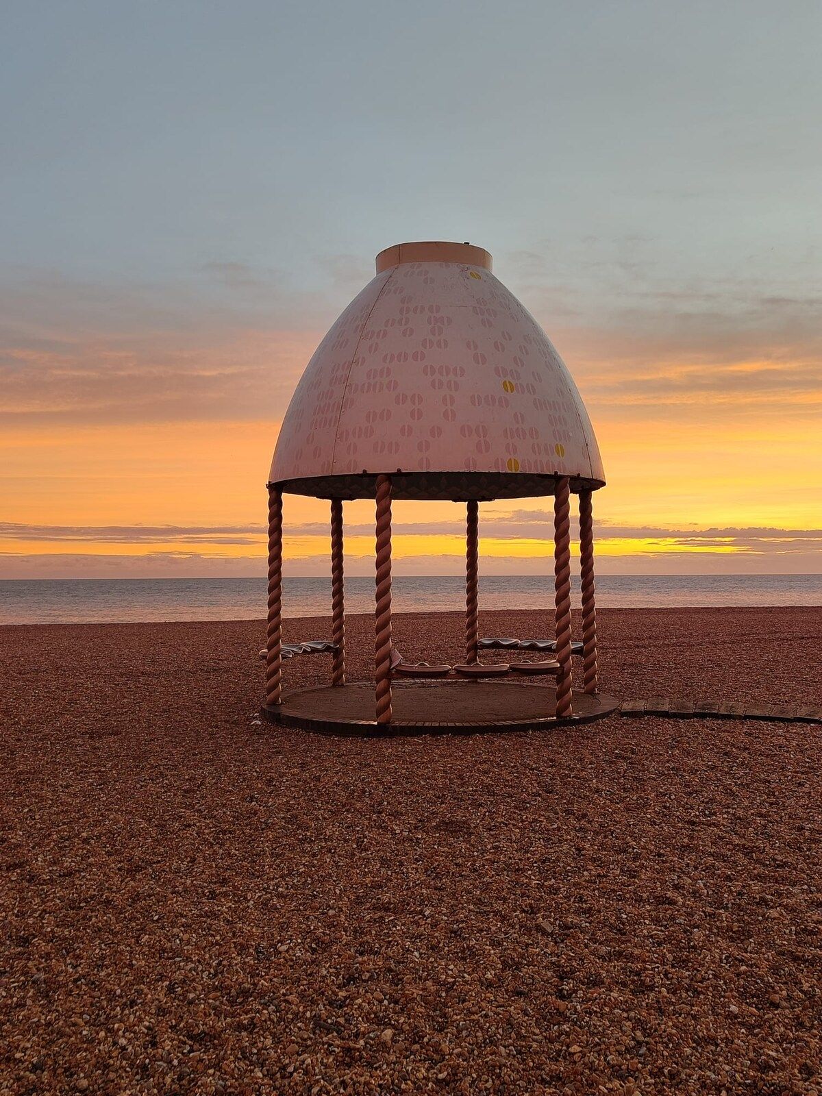 Un quiosco con techo abovedado en una playa de guijarros al atardecer.