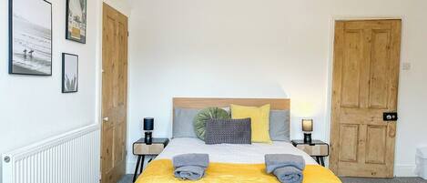 Un dormitorio con una cama, dos mesitas de noche, una puerta de madera y cuadros en la pared.