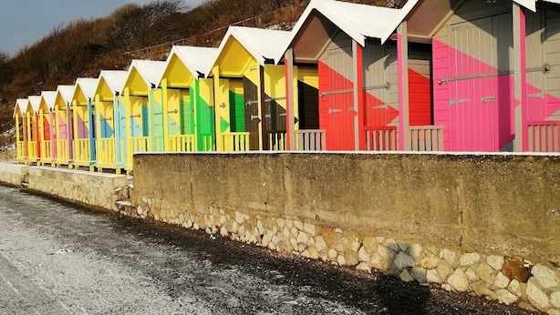 Eine Reihe von bunten Strandhäusern mit weißen Dächern, angeordnet entlang einer Steinmauer neben einer Straße.