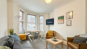Ein modernes Wohnzimmer mit einer Couch, einem Sessel, einem Couchtisch und einem Esstisch. Große Fenster, ein Flachbildfernseher und Kunstwerke an der Wand sind ebenfalls vorhanden.