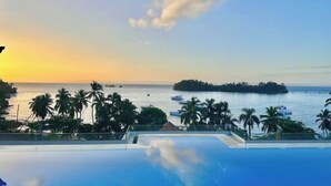 Une vue dégagée sur une piscine, avec un coucher de soleil en arrière-plan, des palmiers et des bateaux sur l’eau.