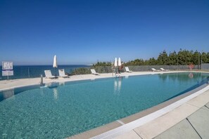 Una piscina azul cristalina con sillas de descanso blancas y sombrillas, una valla y vistas al mar.