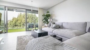 Een moderne woonkamer met een grijze bank, een salontafel en een groot raam met uitzicht op bomen.