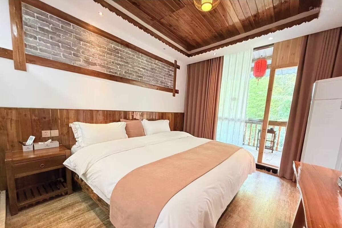 Ein Schlafzimmer mit einem großen Bett, einem Holz-Kopfteil, einem Nachttisch, einer Wand mit Ziegeln und Blick ins Freie.