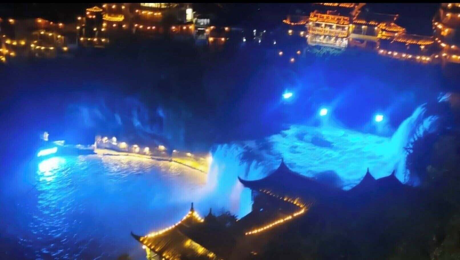 A night view of a water feature with illuminated structures and a traditional building with a curved roof.