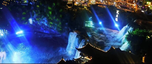 A night view of a waterfall with illuminated structures and a traditional building with a pointed roof. - Tu Wang Ju Ke Zhan (Xiangxi)