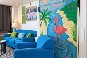 A room with a blue sofa, a painting of a flamingo, and a view of the ocean. - Pinnacle 1471 Oceanfront Tower (Myrtle Beach)