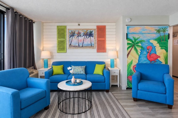 A room with a blue sofa, two armchairs, a round coffee table, and a painting of a beach scene. - Pinnacle 1471 Oceanfront Tower (Myrtle Beach)