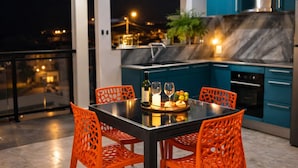 Una cocina moderna con armarios azules y un salpicadero de mármol, un comedor con sillas naranjas y una mesa de cristal, y un balcón con vistas a la ciudad de noche.