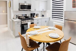 A modern kitchen with white cabinets, a stainless steel refrigerator, and a dining area set for four with a wooden table and brown chairs. - Spacious 3 Bedroom | Steps to Bus to NYC + Views (West New York)