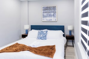 A neatly arranged bedroom with a large bed, two bedside tables, a blue patterned pillow, a brown throw blanket, and a framed artwork on the wall. - Spacious 3 Bedroom | Steps to Bus to NYC + Views (West New York)