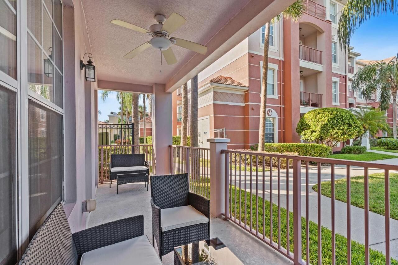 A balcony with wicker furniture and a ceiling fan, overlooking a well-maintained lawn and a building with balconies.