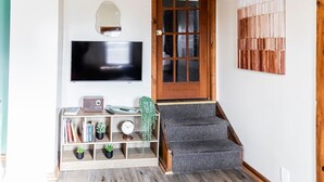 A living room with a TV mounted on the wall, a wooden staircase leading to a door, and a bookshelf with books and plants.