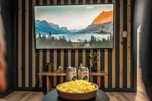 Et værelse med et stort fladskærms-tv monteret på en væg med lodrette træpaneler, et lille træbord med en skål popcorn og en plante.