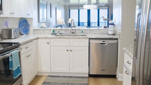 A modern kitchen with white cabinets, stainless steel appliances, a large window, and a view of the ocean. - Barefoot Trace 409 Direct Oceanfront with Private Balcony Overlooking St. Augustine Beach (St. Augustine)