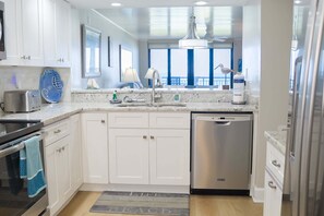 A modern kitchen with white cabinets, stainless steel appliances, a large window, and a view of the ocean. - Barefoot Trace 409 Direct Oceanfront with Private Balcony Overlooking St. Augustine Beach (St. Augustine)