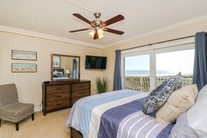 Une chambre avec un lit, une commode, une chaise, un ventilateur de plafond, une télévision et un balcon avec vue sur l’océan.