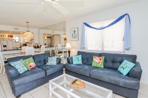 A living room with a dark blue sofa set, a glass coffee table, and a dining area in the background. - Ocean Village Club Condo J26 (St. Augustine)