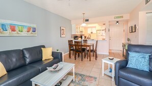 Ein Wohnzimmer mit einem schwarzen Ledersofa, einem Couchtisch und einem Essbereich mit Holzsesseln. Im Hintergrund ist eine Küche zu sehen.