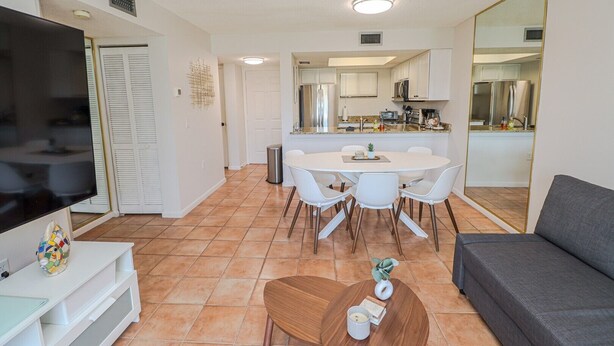 Een moderne woonkamer met een flatscreen televisie, een grijze bank, een eethoek met een witte tafel en stoelen, en een keuken die door een open deur zichtbaar is.