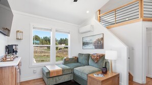 A modern living room with a sectional sofa, a coffee machine, a lamp, and a view of a green landscape. - Huckleberry Inn - 1 BD Cabin (Custer)