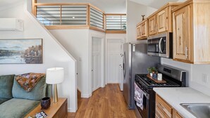 A modern kitchen with wooden cabinets, a stainless steel refrigerator, and a black stove. There is a dining area with a green sofa and a lamp. A staircase leads to an upper level with a glass railing. - Huckleberry Inn - 1 BD Cabin (Custer)