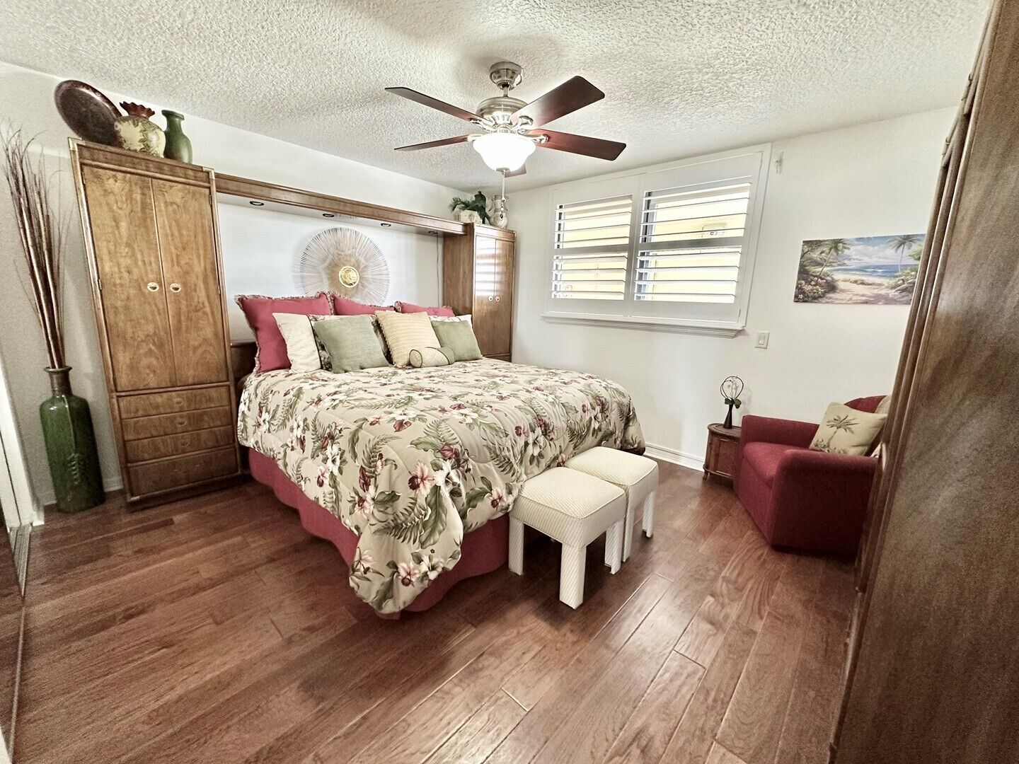 A bedroom with a wooden floor, a bed with floral bedding, a ceiling fan, a window with blinds, and a red armchair.