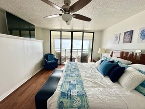 A hotel room with a large bed, a ceiling fan, a view of the ocean through a sliding door, and a chair by the door. - Sand Dollar 3-304 3BR Direct Oceanfront (St. Augustine)