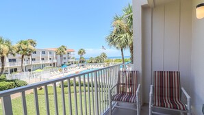 Ein Balkon mit zwei gestreiften Stühlen, ein Poolbereich mit Liegestühlen und Blick auf ein Gebäude und Palmen.