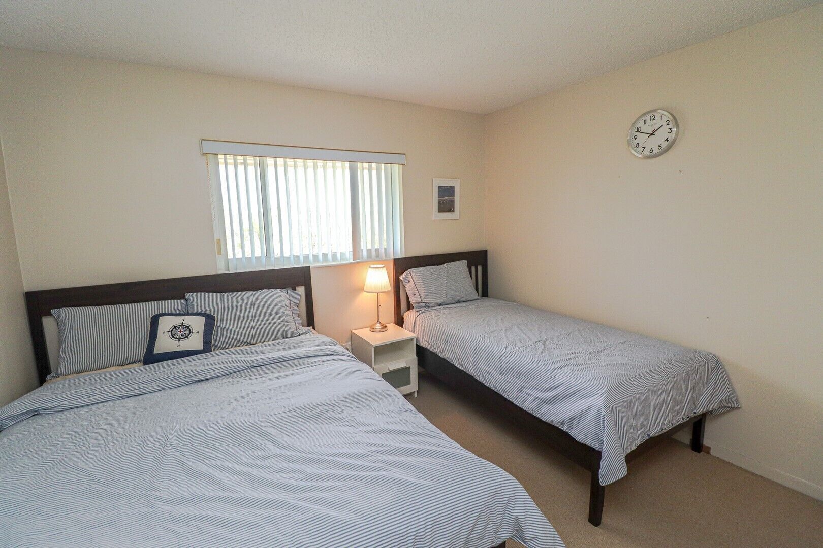 Une chambre avec deux lits simples, une table de chevet avec une lampe, une horloge au mur et une fenêtre avec des stores.