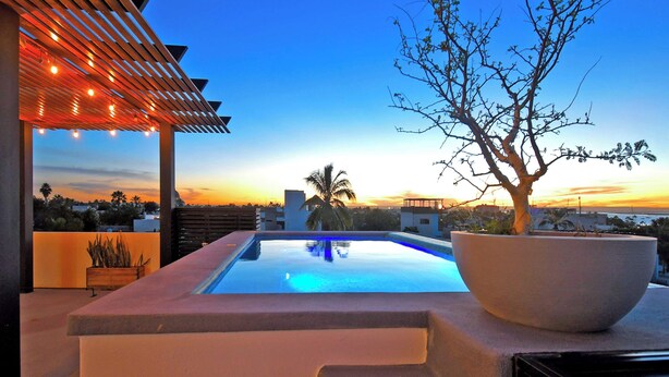 Uma área moderna de piscina externa com pérgola e luzes de corda, um vaso grande e vista para o pôr do sol.