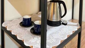 Une étagère métallique à plusieurs niveaux, avec un panier en osier contenant des plantes, des couvertures pliées et une bouilloire en acier inoxydable sur l’étagère inférieure.