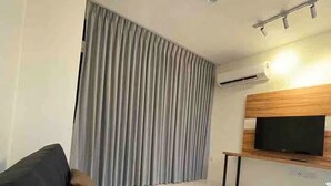 Un salon moderne avec un canapé noir, un rideau gris, un téléviseur fixé au mur et un meuble TV en bois.