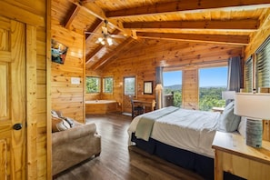 A wooden cabin interior with a bed, a desk, a chair, and a window overlooking a forest. - Epic View | Family Cabin | Hot Tub + Fire Pit | Sleeps 12 | Near Pigeon Forge (Sevierville)