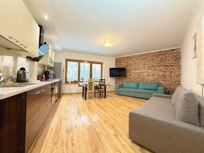 A modern kitchen with wooden flooring, a dining area with a table and chairs, a grey sofa, and a brick wall with a TV mounted above it. - Rynek Wrocław Apartment for 8 People (Wrocław)