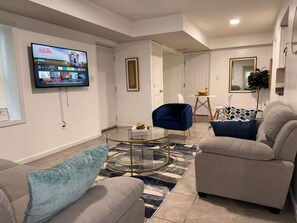 A modern living room with a flat-screen TV mounted on the wall, a glass coffee table, and two sofas with cushions. - Modern Quiet Suite (Newark)