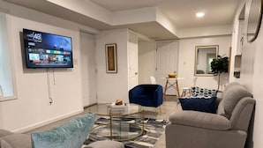 A modern living room with a flat-screen TV mounted on the wall, a glass coffee table, two sofas, and a dining area with a small table and chairs. - Modern Quiet Suite (Newark)