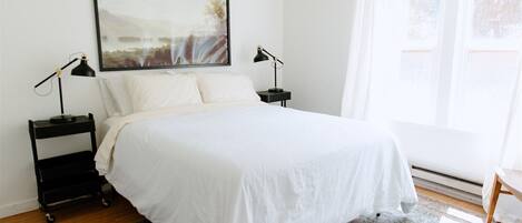 Un lit bien fait, avec du linge de lit blanc, deux tables de chevet avec des lampes, une grande œuvre d’art encadrée au-dessus du lit et une fenêtre avec des rideaux légers.