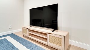 Uma TV de tela plana sobre um móvel de madeira com prateleiras e um painel lateral de vime, colocada em um quarto com um tapete listrado em azul e branco.
