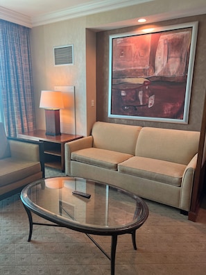 A hotel room with a sofa, a glass coffee table, a lamp, and a large abstract painting on the wall.